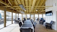 Located at 1015 Nutt Street in Wilmington, NC, this single-story waterfront space offers a captivating blend of rustic craftsmanship and open-air design along the Cape Fear River. The structure is defined by its impressive exposed wood timber framing and beam ceiling, which lend a warm, pavilion-like character to the interior. Large open-air openings line the perimeter, flooding the space with natural light and providing panoramic views of the surrounding marina and waterway. A generous communal dining table, flanked by wicker lounge chairs, serves as the centerpiece of the main gathering area, making it well-suited for group events or casual waterfront dining. Pendant lighting hangs in clusters from the timber ceiling, adding an industrial yet inviting touch to the atmosphere. The light-toned wood plank flooring complements the natural palette of the structure throughout. Additional seating areas near the open edges of the space allow guests to take full advantage of the outdoor scenery. The overall ambiance is relaxed and airy, with the sounds and sights of the waterfront seamlessly integrated into the experience, making this a distinctive and memorable destination on the Wilmington riverfront.