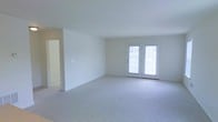 This single-floor space offers a clean, move-in-ready layout defined by its bright and airy atmosphere. The main living area is generously proportioned, featuring wall-to-wall plush carpeting in a light neutral tone that adds warmth and comfort underfoot. Smooth white walls and a white ceiling create a crisp, open canvas well-suited for a variety of décor styles. A standout feature of the living area is the classic French door set, which fills the room with an abundance of natural light while providing direct access to the outdoors. The doors are framed simply, complementing the understated, functional aesthetic of the space. A hallway visible along one side of the living area suggests additional rooms beyond, offering a logical and practical flow throughout the floor plan. The overall ambiance is calm and neutral, making it a versatile space ready to be personalized. The combination of ample natural light, open layout, and soft carpeting gives this space a welcoming and comfortable character suitable for everyday living.