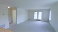 This single-floor space offers a clean, move-in-ready layout defined by its bright and airy atmosphere. The main living area is generously proportioned, featuring wall-to-wall plush carpeting in a light neutral tone that adds warmth and comfort underfoot. Smooth white walls and a white ceiling create a crisp, open canvas well-suited for a variety of décor styles. A standout feature of the living area is the classic French door set, which fills the room with an abundance of natural light while providing direct access to the outdoors. The doors are framed simply, complementing the understated, functional aesthetic of the space. A hallway visible along one side of the living area suggests additional rooms beyond, offering a logical and practical flow throughout the floor plan. The overall ambiance is calm and neutral, making it a versatile space ready to be personalized. The combination of ample natural light, open layout, and soft carpeting gives this space a welcoming and comfortable character suitable for everyday living.