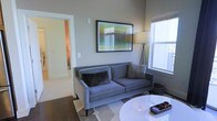 This single-floor space presents a thoughtfully designed living area that blends contemporary style with a functional layout. The living room serves as the centerpiece, featuring neutral gray walls that create a calm, sophisticated backdrop. A plush gray sofa anchors the seating area, complemented by decorative throw pillows and positioned atop a patterned area rug that adds texture to the dark hardwood flooring beneath. A framed abstract artwork with vibrant color accents hangs prominently above the sofa, adding a modern artistic touch to the space. A sleek white round coffee table sits in the foreground, contributing to the room's clean, minimalist aesthetic. Natural light filters in through a large window fitted with a shade, brightening the room and enhancing its open feel. A tripod floor lamp positioned beside the window provides additional ambient lighting. A wide doorway visible from the living area leads to an adjacent hallway, hinting at further rooms beyond. The overall ambiance is modern and inviting, with cohesive design choices that give the space a polished, well-curated appearance.