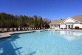 This inviting outdoor amenity space is designed for relaxation and recreation, centered around a generously sized, freeform swimming pool with a smooth surrounding deck lined with lounge chairs on both sides. The pool's clear, turquoise water reflects the open sky, creating a resort-like atmosphere. A classic clubhouse with a pitched roof, chimney, and large windows serves as the architectural backdrop, reinforcing the welcoming character of the community.

A substantial pergola structure anchors one side of the space, providing shaded seating beneath its open-lattice roof. Beneath it, comfortable wicker furniture with vibrant teal cushions — including a loveseat, armchair, and coffee table — creates a cozy outdoor living area. Adjacent to the pergola is a well-equipped outdoor kitchen station featuring a built-in gas grill and ample counter space, ideal for al fresco dining and entertaining.

The concrete patio extends throughout, offering additional seating areas with patio chairs and a table shaded by a teal market umbrella. Wrought iron fencing encloses the perimeter, ensuring privacy and safety. Beyond the fence, a lush backdrop of mature pine and deciduous trees provides natural screening and a peaceful, wooded ambiance that enhances the overall outdoor experience throughout every season.