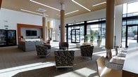 This contemporary lobby is designed to make a strong first impression through a combination of striking architectural elements and carefully curated furnishings. The space features soaring double-height ceilings that create a grand sense of openness, supported by cylindrical columns with warm wood-toned finishes that add a natural contrast to the crisp white walls.

Bold burnt-orange accent walls serve as a defining design feature, lending the space a vibrant, energetic character that is echoed throughout the furnishings, including an orange accent chair and coordinating throw pillows. One lounge area offers a comfortable sectional sofa paired with a light wood coffee table adorned with potted greenery and decorative objects, while framed artwork on the orange walls adds a curated, gallery-like touch.

The opposite end of the lobby opens up dramatically with floor-to-ceiling glass doors and windows, inviting abundant natural light and providing clear sightlines to the surrounding streetscape. This area is furnished with a cluster of patterned upholstered armchairs arranged around a central coffee table, creating an inviting gathering space. Linear recessed lighting and a warm wood-paneled ceiling detail enhance the modern aesthetic. A reception or service counter is visible along one wall, alongside a wall-mounted display screen, completing the functional layout of this well-appointed lobby.