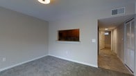 This single-floor space offers a practical and straightforward layout designed for comfortable everyday living. The main living area is finished with wall-to-wall gray carpeting, which adds a soft, neutral foundation to the room. Walls are painted in a clean, light gray tone throughout, lending the space a cohesive and understated aesthetic. A flush-mount ceiling light fixture provides overhead illumination in the living area.

A distinctive architectural feature is the pass-through window cut into the wall between the living area and the kitchen. This opening is framed with a white ledge that doubles as a small counter or serving surface, promoting a sense of connectivity between the two spaces. Through the pass-through, warm-toned, medium-brown cabinetry is visible along with a dark tile backsplash and a sink, suggesting a well-appointed kitchen.

Adjacent to the living area, a hallway leads further into the space, where tile flooring transitions from the carpeted zone, and a door with a light interior is visible — likely leading to a bathroom or secondary room. The overall ambiance is clean, neutral, and move-in ready, with a simple, functional design throughout.