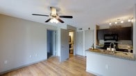 This single-floor space features an open-concept layout that connects the living area and kitchen, creating a cohesive and functional flow throughout. The living area is finished with warm wood-style laminate flooring and neutral gray walls, contributing to a clean, modern aesthetic. A ceiling fan with a built-in light fixture is centrally mounted, providing both air circulation and ambient lighting.

The kitchen is positioned along one side of the space and is equipped with dark espresso-toned cabinetry, a granite or stone-look countertop, and a built-in range with overhead microwave. A peninsula-style counter extends into the living space, offering additional prep area and casual seating potential.

Adjacent to the living area, two doors lead to additional rooms — one opening to a full bathroom featuring a toilet, vanity, and mirror, and the other leading to what appears to be a bedroom or closet. A separate closet door is also visible nearby. The textured ceiling adds a classic residential finish to the space, while HVAC venting ensures climate control throughout. Overall, the space presents a practical and tidy interior suited for everyday living.