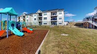 Located at 9 Monticello Dr in Brewster Village, NY, this thoughtfully designed outdoor amenity space serves as a central hub for recreation and community engagement. The grounds are characterized by well-maintained open green lawns that create a spacious, welcoming atmosphere beneath wide-open skies. A colorful, multi-feature playground set on a rubber mulch safety surface provides a dedicated area for children, complete with slides and climbing structures. Adjacent to the playground, fenced sports courts offer space for active recreation, while picnic tables and seating areas are strategically placed throughout the grounds for gathering and relaxation. Smooth concrete walking paths wind through the property, connecting the various amenity zones and encouraging outdoor movement. A standout architectural feature is the large clubhouse building, designed in a modern farmhouse style with crisp white board-and-batten siding, a metal roof, expansive black-framed windows, and an elevated wraparound deck accessible by exterior staircases. Surrounding residential buildings, finished in neutral siding with private balconies, frame the green space and contribute to the cohesive community aesthetic. The combination of recreational facilities, open landscaping, and well-considered pathways makes this outdoor space a comprehensive and functional amenity for residents.
