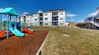 Located at 9 Monticello Dr in Brewster Village, NY, this thoughtfully designed outdoor amenity space serves as a central hub for recreation and community engagement. The grounds are characterized by well-maintained open green lawns that create a spacious, welcoming atmosphere beneath wide-open skies. A colorful, multi-feature playground set on a rubber mulch safety surface provides a dedicated area for children, complete with slides and climbing structures. Adjacent to the playground, fenced sports courts offer space for active recreation, while picnic tables and seating areas are strategically placed throughout the grounds for gathering and relaxation. Smooth concrete walking paths wind through the property, connecting the various amenity zones and encouraging outdoor movement. A standout architectural feature is the large clubhouse building, designed in a modern farmhouse style with crisp white board-and-batten siding, a metal roof, expansive black-framed windows, and an elevated wraparound deck accessible by exterior staircases. Surrounding residential buildings, finished in neutral siding with private balconies, frame the green space and contribute to the cohesive community aesthetic. The combination of recreational facilities, open landscaping, and well-considered pathways makes this outdoor space a comprehensive and functional amenity for residents.
