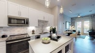 This thoughtfully designed single-floor residence showcases a cohesive modern aesthetic carried throughout every room. The open-concept kitchen is a standout feature, outfitted with crisp white shaker cabinetry, quartz countertops, a decorative marble-patterned tile backsplash, and a full suite of stainless steel appliances including a French door refrigerator, range, microwave, and built-in wine cooler. A central kitchen island provides additional prep space and casual seating, while pendant lighting adds a refined touch overhead. The living area impresses with soaring ceilings and floor-to-ceiling glass doors that flood the room with natural light and open onto a private balcony with outdoor views. Light wood-style flooring runs through the main living spaces, complementing the neutral color palette. The primary bedroom features plush carpet, a bold teal accent wall, and large windows offering outdoor views, while the secondary bedroom is equally well-lit with generous window placement. A hallway connects the bedrooms to a full bathroom finished with white cabinetry, a quartz-topped vanity, wood-look tile flooring, and a tub-shower combination. The layout flows efficiently, making excellent use of the available space throughout.