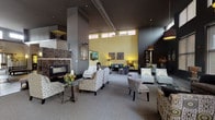 This single-story community amenity space is thoughtfully designed with a sophisticated palette of dark charcoal, warm yellow accents, and neutral tones throughout. The grand entrance lobby welcomes visitors with soaring ceilings, wood-look flooring, large clerestory windows that draw in natural light, and carefully curated décor including a decorative glass feature wall and elegant console furnishings.

The expansive main lounge centers around a striking floor-to-ceiling marble-tiled fireplace, anchoring multiple seating arrangements with patterned armchairs and upholstered furniture. Adjacent to this area, a darker-toned lounge offers oversized windows with exterior views, creating an intimate yet well-lit gathering space with glass-top coffee tables and richly textured seating.

A separate sitting area features two grey sofas accented with teal throw pillows, complemented by a cozy dining nook with a natural wood table and upholstered chairs in mixed tones. Throughout, pendant lighting and wall sconces provide warm, layered illumination.

The fitness center is well-equipped with cardio machines including ellipticals, treadmills, and stationary bikes, alongside strength training equipment, free weights, and wall mirrors, making it a comprehensive facility for residents.