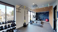 This contemporary fitness space is designed to support a full range of workout routines within a compact, efficiently organized layout. The room benefits from abundant natural light, courtesy of large windows along the perimeter walls that offer views to the outdoors and contribute to an open, energizing atmosphere. The ceiling features a combination of recessed lighting and a mounted HVAC unit, complemented by warm wood slat accents that add a refined, modern touch to the space.

Equipment is thoughtfully arranged throughout the room. A wall-mounted pull-up and suspension bar system with TRX straps anchors one side of the space, ideal for bodyweight and functional training. A dumbbell rack lines the adjacent wall, providing a range of free weight options. Toward the rear of the room, rowing machines are positioned on a rubberized mat area, suitable for low-impact cardio training.

A bold, typographic mural spanning one full wall displays motivational words and phrases, reinforcing a focused and driven atmosphere. The flooring throughout is durable and gym-appropriate, designed to withstand heavy use. Overall, the space strikes a balance between functionality and modern design.