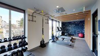 This contemporary fitness space is designed to support a full range of workout routines within a compact, efficiently organized layout. The room benefits from abundant natural light, courtesy of large windows along the perimeter walls that offer views to the outdoors and contribute to an open, energizing atmosphere. The ceiling features a combination of recessed lighting and a mounted HVAC unit, complemented by warm wood slat accents that add a refined, modern touch to the space.

Equipment is thoughtfully arranged throughout the room. A wall-mounted pull-up and suspension bar system with TRX straps anchors one side of the space, ideal for bodyweight and functional training. A dumbbell rack lines the adjacent wall, providing a range of free weight options. Toward the rear of the room, rowing machines are positioned on a rubberized mat area, suitable for low-impact cardio training.

A bold, typographic mural spanning one full wall displays motivational words and phrases, reinforcing a focused and driven atmosphere. The flooring throughout is durable and gym-appropriate, designed to withstand heavy use. Overall, the space strikes a balance between functionality and modern design.
