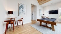 Located at 3788 Old Port Royal Rd N in Spring Hill, Tennessee, this single-floor living room is a striking example of contemporary design. The space is defined by its dramatic vaulted ceiling accented with warm wood paneling, creating an airy, open atmosphere that feels notably expansive. Light-toned hardwood flooring runs throughout, lending a clean, cohesive foundation to the room.

A standout architectural feature is the floor-to-ceiling multi-pane window wall framed in bold dark metal, which floods the interior with abundant natural light and offers unobstructed views of the surrounding landscape and greenery. This feature serves as a dramatic focal point and seamlessly connects the indoors to the outdoors.

The living area is thoughtfully appointed, centered around a substantial wood-framed billiards table with a navy felt surface, positioned atop a soft area rug. A bar-height table with warm wood stools provides a casual seating vignette, while curated abstract artwork adorns the crisp white walls, adding a refined artistic touch. Recessed lighting complements the natural light throughout.

An open passage connects this space to an adjacent dining and kitchen area, enhancing the overall flow of the home. The overall ambiance is modern, bright, and inviting — ideal for both relaxed entertaining and everyday living.