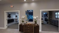 This single-floor space makes a strong first impression with a thoughtfully designed entryway that sets the tone for the rest of the interior. The central foyer features a dark wood console table adorned with white orchids and decorative accents, anchored by a bold blue and gold abstract artwork mounted on a neutral gray wall. Recessed lighting highlights the curated arrangement, drawing the eye through two open doorways that lead into adjacent rooms.

To the left, a dining area is visible, outfitted with modern furnishings and benefiting from natural light filtered through a nearby window. The clean lines and contemporary aesthetic carry throughout this space.

To the right, a spacious living area showcases a striking stone fireplace that serves as a natural focal point, complemented by soft, neutral upholstery and warm ambient lighting. The stone surround adds a textural, organic element that contrasts beautifully with the otherwise modern interior.

Throughout the space, the design palette remains cohesive — warm neutrals, rich wood tones, and carefully chosen decorative elements contribute to an atmosphere that is both refined and welcoming. The open layout encourages a natural flow between spaces, making the interior feel connected and expansive while maintaining distinct functional areas.