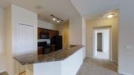 This single-floor residence offers a thoughtfully designed open-concept layout that maximizes both functionality and comfort. The kitchen serves as a focal point of the space, showcasing rich dark wood cabinetry, granite countertops, and a full suite of black appliances, including a refrigerator, electric range, and built-in microwave. A generously sized peninsula counter extends into the living area, creating a natural divide between the kitchen and the adjacent carpeted living space while offering additional seating and prep area. Track lighting above the kitchen provides focused illumination, complementing the warm neutral tones found throughout. The soft carpeting in the main living area adds a comfortable, inviting quality to the space. A hallway visible from the main area leads to additional rooms, suggesting a well-organized floor plan with clear separation between living and private spaces. Closet space is evident along the corridor, adding to the home's practical storage solutions. The overall ambiance is warm and functional, with a clean, move-in-ready aesthetic defined by consistent neutral wall tones and a cohesive design throughout the single level.