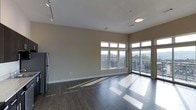 This single-floor space is defined by its open-concept layout and clean, modern design. The main living area features rich, dark hardwood flooring that contrasts elegantly with the neutral-toned walls, creating a polished and contemporary atmosphere. Notably tall ceilings amplify the sense of spaciousness, while a combination of track lighting and a flush-mount ceiling fixture provide versatile illumination throughout the space.

Large, multi-pane windows line the exterior wall, flooding the interior with abundant natural light and framing expansive views of the surrounding urban skyline. A sliding glass door provides direct access to an outdoor balcony, further connecting the interior to the city environment beyond. The generous window placement allows sunlight to cast dramatic patterns across the hardwood floors, enhancing the open, airy feel of the room.

A stainless steel refrigerator is visible near the perimeter, hinting at an adjacent kitchen area. The overall design aesthetic is minimalist and contemporary, with clean lines, neutral finishes, and an emphasis on natural light and urban views as central design features.