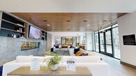 Located at 308 NJ-31 N in Flemington, New Jersey, this single-floor community lounge offers a generously sized, open-plan layout designed with a contemporary aesthetic and thoughtful attention to detail. The space is anchored by a striking marble-clad, see-through fireplace flanked by built-in shelving, creating a natural focal point within the main living area. Overhead, warm wood-slatted ceiling panels add texture and sophistication, complementing the light hardwood flooring that runs throughout. Large floor-to-ceiling glass entry doors and an array of oversized windows flood the interior with natural light while providing views of the surrounding residential community and tree-lined landscape. The lounge is divided into distinct yet connected zones. A plush central seating arrangement features a generous sectional sofa and coordinating accent chairs, ideal for social gatherings. A separate intimate nook is furnished with rounded navy velvet lounge chairs and a brushed gold accent table beneath statement pendant lighting. A dining area with leather chairs surrounds a round table set against a bold geometric-patterned feature wall. An additional open area with a wall-mounted display screen offers flexible use. The overall ambiance is polished, welcoming, and well-appointed throughout.