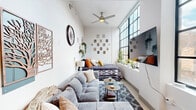 Located at 128 Sixth St E in downtown Cincinnati, Ohio, this single-floor studio makes a striking impression through clever design and efficient use of space. The living area is defined by dramatic soaring ceilings and a generous run of floor-to-ceiling industrial-style windows that flood the room with natural light. A ceiling fan adds to the airy feel, while the open layout comfortably accommodates both a seating and sleeping area side by side, giving the space a functional yet cozy loft-like ambiance.

The galley kitchen is sleek and well-appointed, featuring white cabinetry, a tile backsplash, light hardwood-style flooring, stainless steel appliances including a refrigerator and range, and ample counter space along both sides. Open shelving adds a modern touch for displaying everyday essentials.

A hallway connects the main living space to the bathroom, where exposed brick detailing adds warmth and character. The bathroom is clean and contemporary, with a fully tiled glass-enclosed walk-in shower, open wooden shelving, and bright white finishes throughout.

Throughout the studio, white walls provide a neutral backdrop that enhances the sense of openness, while thoughtful décor and greenery create a welcoming, lived-in atmosphere. This is a well-optimized urban retreat in the heart of Cincinnati.