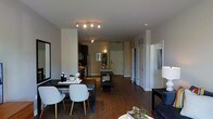 This single-floor residence showcases an open-concept design that connects the living, dining, and kitchen areas into one cohesive space. Dark hardwood flooring runs throughout, providing a warm, grounding contrast to the light grey walls and white trim. The dining area features a sleek dark table paired with modern light grey chairs, positioned beneath a contemporary track lighting fixture that illuminates the space effectively. Adjacent to the dining area, the kitchen is equipped with stainless steel appliances, including a refrigerator and range, set against dark cabinetry. A small utility cart serves as additional workspace or storage between the kitchen and dining zones. Toward the rear of the space, a neutral-toned entryway leads to what appears to be a front door, flanked by a built-in shelving unit. Multiple white paneled doors line the hallway, likely providing access to additional rooms or storage. A partial view of the living area to the right reveals a dark sofa and side table. The overall ambiance is modern and functional, combining practical design elements with a cohesive, contemporary aesthetic throughout.