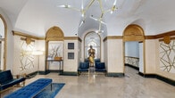 This distinguished lobby space makes an immediate architectural statement with its soaring groin-vaulted ceilings, gracefully framed by gold-trimmed arched alcoves lining the perimeter walls. The flooring is composed of polished marble tiles that reflect the abundant light from a sculptural gold branching chandelier suspended overhead, reinforcing the space's refined aesthetic.

The main seating area is furnished with tufted royal blue velvet benches and gold-framed accent chairs, anchored by a richly toned blue area rug that adds warmth and contrast against the light marble floors. A striking full-length arched mirror amplifies the sense of depth and openness throughout.

A centerpiece armored knight statue stands prominently beneath a stained glass window, lending a dramatic, museum-like quality to the interior. Decorative wall art and floral arrangements are tastefully positioned on a marble-topped console table, contributing to the curated atmosphere.

The space extends into a connecting corridor featuring bold geometric gold line patterns across the walls, a vintage-style letter box, elevator access, and a resident mailbox panel — blending historic character with modern functionality. Glass entry doors at the street-facing end flood the foyer with natural light, providing a seamless visual connection to the exterior.