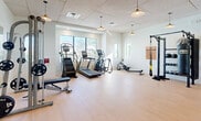 Located in Bonita Springs, Florida, this fitness center offers a comprehensive workout facility designed to support a wide range of exercise routines. The space features light hardwood-style flooring throughout, crisp white walls, and a drop ceiling fitted with stylish pendant lights that provide warm, even illumination across the open floor plan.

Natural light flows in generously through multiple large windows, offering views of the surrounding Florida landscape and creating a bright, energizing atmosphere. A full-length mirror wall on one side of the room visually expands the space and aids in form monitoring during workouts.

The cardio zone is well-appointed with treadmills, elliptical trainers, a stair climber, and a rowing machine. The strength training area includes a multi-functional cable machine, a power rack with TRX suspension straps, a punching bag, and a dedicated free weight section featuring a full dumbbell rack with an adjustable bench. Kettlebells, medicine balls, and a stability ball are also available for functional and cross-training exercises.

The layout provides ample open floor space between equipment stations, allowing for comfortable movement and functional training. Access is provided through a glass-panel door, and the facility is clean, modern, and thoughtfully organized to accommodate multiple users simultaneously.