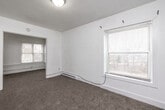 Located at 1042 18th St in Des Moines, Iowa, this 2-bedroom, 1-bathroom unit offers a practical, single-floor layout designed for efficient everyday living. The space is characterized by clean white walls, neutral carpeting throughout the living and bedroom areas, and classic white baseboards that lend a simple, uniform aesthetic.

The layout includes two distinct living room areas, providing flexibility for separate lounging and dining arrangements. Natural light filters in through multiple windows fitted with blinds, giving the space a bright and open feel. Baseboard heaters run along the walls in several rooms, ensuring consistent warmth throughout the colder months.

The kitchen features wood-look vinyl plank flooring, contrasting with the carpeted areas elsewhere. It is equipped with a full-size refrigerator and a freestanding electric range, along with upper cabinetry for storage.

Both bedrooms are carpeted and offer dedicated closet access, with the primary bedroom providing a comfortable footprint for standard furnishings. The second bedroom also benefits from natural light via a window with exterior views.

Two closets serve the unit's storage needs, and the overall layout flows logically from room to room. The white interior palette and straightforward design make this a clean, move-in ready space in a residential Des Moines neighborhood.