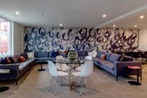 Located at 868 S Fifth St in San Jose, CA, this single-floor communal lounge is a thoughtfully designed gathering space that blends contemporary aesthetics with functional versatility. The room's most striking feature is a large-scale, abstract mural that spans an entire accent wall, rendered in sweeping brushstroke patterns of deep navy and charcoal against a textured neutral backdrop, immediately drawing the eye and setting an artistic tone throughout the space. Generous sectional sofas in soft grey, accented with vibrant orange, navy, and cream throw pillows, are arranged to encourage both intimate conversation and larger group gatherings. A round glass-top dining table with sleek white modern chairs provides a casual dining or collaborative work area, while additional round tables with black molded chairs near the mural wall offer further seating flexibility. A built-in counter-height bar ledge with black leather stools lines a perforated metal railing, offering additional seating with a view into the level below. Large windows along the perimeter flood the space with natural light and frame views of the surrounding San Jose neighborhood. Recessed ceiling lighting and light-toned carpet flooring complete the clean, modern interior, making this lounge an inviting and multifunctional communal retreat.