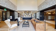 Located at 3750 Chamberlain Lane in Louisville, Kentucky, the leasing office and amenity hub of The Louis Apartment Homes is a single-story space designed with a contemporary yet warm aesthetic. The grand entrance greets visitors with floor-to-ceiling steel-framed glass doors and windows that flood the interior with natural light, complemented by wide-plank hardwood flooring throughout.

The main lobby is defined by dramatic soaring ceilings and two imposing natural stone accent walls that anchor the space on either side, lending a refined, upscale character. Large sliding barn doors with black steel and glass paneling provide a stylish transition between zones. A centrally positioned leasing desk features a dark paneled base with a crisp white countertop, illuminated by industrial-style pendant lights overhead.

The seating area is furnished with a leather sofa, modern accent chairs, and a woven area rug, creating a comfortable and approachable lounge atmosphere. A dedicated mail room offers built-in mailboxes, a work counter with under-cabinet storage, and task lighting. Adjacent to the lobby, a coworking lounge provides a dining-height marble-top table with pendant lighting, a coffee station, and glass-enclosed workspaces with views to the outdoors, offering a functional and design-forward environment for residents.