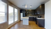 This single-floor space showcases a thoughtfully designed open-concept layout centered around a contemporary kitchen. Dark espresso-toned cabinetry lines the walls, providing generous storage alongside a dedicated built-in wine rack that adds a refined touch. The kitchen is equipped with stainless steel appliances, including a refrigerator, electric range, and microwave, complemented by a mosaic tile backsplash that introduces subtle texture and visual interest. A central island with a clean white countertop serves as both a functional workspace and a natural gathering point, illuminated overhead by three stylish black pendant lights that anchor the space with a modern aesthetic. Warm-toned hardwood flooring flows throughout, creating a cohesive and inviting atmosphere. Large floor-to-ceiling windows fitted with horizontal blinds flood the area with natural light while offering views of surrounding greenery. Recessed lighting and a ceiling-mounted track light provide additional illumination throughout the kitchen area. The overall design balances contemporary finishes with a warm, functional ambiance.