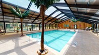 This indoor aquatic facility is designed to evoke a resort-like atmosphere year-round. The centerpiece is a generously sized rectangular swimming pool with crystal-clear blue water, accompanied by a separate hot tub, both set within a spacious pool deck finished in textured aggregate concrete. Decorative artificial palm trees are strategically placed throughout, enhancing the tropical ambiance.

The architectural highlight of the space is its dramatic pitched roof structure, composed of large translucent and glass panels supported by a bold steel framework. This design allows abundant natural light to pour in from above, creating a bright and airy environment throughout the day. Floor-to-ceiling glass walls and sliding glass doors line the perimeter, offering views of the surrounding outdoor grounds while maintaining a fully enclosed, climate-controlled interior.

The pool deck is well-appointed with lounge chairs, seating areas, and small tables, providing ample space for relaxation. One end of the facility features warm cedar-paneled walls, lending a natural, rustic character to the space. Safety equipment, potted tropical plants, and thoughtful signage complete the interior. The single-level layout ensures easy accessibility and a seamless flow throughout the entire facility.