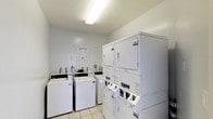 This single-floor shared laundry facility is designed with practicality and efficiency in mind. The room features a clean, straightforward layout with light-colored walls and a bright fluorescent overhead light that ensures ample visibility throughout the space. The flooring consists of easy-to-maintain light-toned tiles, contributing to the room's tidy and functional atmosphere. Multiple top-loading, coin-operated washing machines are lined along one wall, each equipped with individual controls and payment mechanisms. Complementing the washers, a bank of stacked coin-operated dryers is positioned along the adjacent wall, maximizing the use of vertical space and increasing the overall laundry capacity of the room. Instructional and operational signage is posted on the wall, providing users with clear guidance on how to properly operate the machines. The compact yet well-organized design of this laundry room makes it a convenient and accessible amenity, offering residents a reliable and straightforward space to manage their laundry needs on a single level.