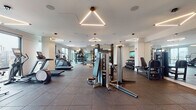 Located at 4601 N Broadway St in Chicago's Uptown neighborhood, this contemporary fitness center is a well-appointed, single-floor facility designed to support a comprehensive workout experience. The space is defined by its clean, modern aesthetic, featuring crisp white walls, rubber-tiled flooring, and striking geometric triangular pendant lighting that adds a distinctive architectural character throughout.

The cardio zone is positioned along a wall of large floor-to-ceiling windows, offering panoramic views of the Chicago skyline — an energizing backdrop for treadmills, ellipticals, stair climbers, and a stationary bike. Natural light floods this area, creating a bright and motivating environment.

The strength training section is generously equipped with a multi-functional cable and power rack system, a full dumbbell rack, weight benches, rowing machines, and leg press equipment. Floor-to-ceiling mirrors line key areas of the space, enhancing the sense of openness while allowing users to monitor their form.

A dedicated functional training area provides space for stability balls and accessories, while a separate entry vestibule features a water fountain and access to an adjoining sauna room with warm wood-paneled walls. The overall ambiance is polished and professional, combining high-end equipment with thoughtful design to create an inviting, fully functional fitness facility.