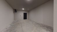 This single-level garage offers a generously proportioned, open layout designed for practical utility. The space is finished with smooth, neutral-toned drywall on all walls and ceiling, giving it a clean, well-maintained appearance uncommon in standard garages. The concrete slab floor spans the full length of the space, providing a durable, easy-to-maintain surface suitable for vehicle storage, workshop use, or additional storage needs.

A motorized sectional roll-up garage door is positioned at one end, equipped with an automatic opener unit mounted to the ceiling, ensuring convenient vehicle access. Recessed overhead lighting provides adequate illumination throughout the interior. A standard interior access door with a dark-finished frame is located on the far wall, offering direct entry to an adjacent interior space.

The elongated footprint of the garage, clearly visible in the overhead layout view, provides ample room to accommodate multiple vehicles or serve a variety of alternative uses. The neutral color palette, finished walls, and clean construction details contribute to a polished, move-in-ready environment. Overall, the space presents a versatile and well-constructed garage suitable for a broad range of functional purposes.
