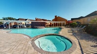 This outdoor amenity space serves as a central recreational hub for its residential community. The centerpiece is a generously sized, freeform swimming pool with striking blue-tiled water, complemented by a circular spa/hot tub positioned at its edge — ideal for relaxation. A standout architectural feature is the brick water feature structure rising from within the pool, adding a decorative focal point to the overall design.

Surrounding the pool is a spacious, stamped concrete deck finished in an earthy tone, providing ample room for lounge seating and circulation. A white pergola-style covered structure runs along one side of the pool area, offering shaded seating and dining space underneath, further enhanced by patio tables, chairs, and umbrellas.

The pool area is enclosed by a black iron security fence with a gated entry, clearly marked with posted pool rules for resident safety. Manicured green lawns and walkways connect the pool enclosure to the surrounding multi-story residential buildings, which feature brick and stucco exteriors with balconies. The entire space benefits from abundant natural light and open skies, creating a bright and inviting atmosphere well-suited for outdoor recreation and community gathering.