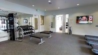 This fitness center is organized across a single floor and divided into distinct functional zones designed to support a comprehensive workout experience. The cardio area is anchored by a row of modern treadmills and an elliptical machine, each equipped with individual screens for entertainment during exercise. A large window along the back wall allows natural light to filter in while offering views of the surrounding outdoor environment. A wall-mounted flat-screen television provides an additional viewing option for users in this zone.

The free weights area is outfitted with a fully stocked dumbbell rack spanning a wide range of weights, along with multiple adjustable flat benches for strength training exercises. Full-length mirrors line the walls, enhancing the sense of space and allowing users to monitor their form. A medicine ball rack and additional training accessories are also present. Recessed ceiling lights throughout both areas provide consistent, even illumination. The flooring consists of durable, dark-toned carpet suitable for high-traffic fitness use. The overall aesthetic is clean and modern, with neutral walls and a well-organized layout that promotes efficient use of the space.