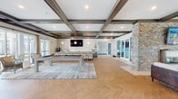 This single-floor community clubhouse is designed with a contemporary yet welcoming aesthetic, blending modern finishes with warm architectural details throughout. The space is anchored by a striking coffered ceiling accented with dark wood beams and recessed lighting, creating an elevated and refined atmosphere.

The main gathering area features herringbone-patterned wood-look flooring and includes a full-size pool table set atop a large area rug, making it an ideal social hub. Pendant globe lights hang above a generously sized central island surrounded by wooden bar stools, while teal-blue cabinetry with brass hardware lines the walls beneath a wall-mounted television — a polished focal point perfect for entertaining.

The kitchen area is well-appointed with a deep sink, ample counter space, and direct access to the outdoors through a glass door that frames views of palm trees and the surrounding community. A natural stone accent wall adds an organic, textural element near the room's glass-paneled sliding doors, which flood the interior with natural light and provide seamless views of the outdoor pool and courtyard.

A quieter lounge nook offers a plush sectional sofa and additional seating, bathed in soft natural light from large windows dressed with sheer curtains. The overall ambiance is bright, social, and thoughtfully designed for both relaxation and community engagement.
