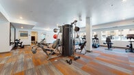 This fitness center in West Haven, Utah offers a comprehensive, single-floor layout designed to support a wide range of workout routines. The main gym area is carpeted in a vibrant multi-toned pattern and houses a variety of strength and cardio equipment, including a True multi-station cable machine, a dumbbell rack with a weight bench, treadmills, elliptical trainers, and stationary bikes. Large windows along the perimeter allow natural light to fill the space and provide views of the surrounding exterior. Full-length mirrors are strategically placed to enhance the sense of space and support proper form during exercise. A dedicated cardio zone features a row of treadmills and step machines positioned in front of windows, creating an open, energizing atmosphere. Adjacent to the main floor is a private spin studio with light wood flooring, spin bikes, and a large wall mirror, offering an ideal setting for cycling workouts. A separate entry area provides convenient towel service and storage. The facility also includes a children's activity room furnished with small tables, stools, bookshelves, and a wall-mounted television, making it a family-friendly amenity. Recessed lighting throughout ensures a bright, well-lit environment across all areas.