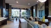 This single-floor space presents a sleek, contemporary design that seamlessly blends indoor comfort with an openness to the outdoors. Expansive floor-to-ceiling windows dominate the exterior-facing walls, flooding the interior with abundant natural light and offering unobstructed views of a lush outdoor courtyard lined with mature trees and greenery. The open-plan layout is thoughtfully arranged with multiple seating areas, including plush gray upholstered armchairs accented with patterned throw pillows and a substantial sofa, creating inviting conversational groupings. A light wood communal table with bar-height seating adds a functional social hub to the space. Recessed lighting in the crisp white ceiling complements the natural light, ensuring a bright and welcoming ambiance throughout. Dark vertical architectural columns and tasteful patterned drapery in gold and gray tones provide striking contrast against the light palette of the walls and flooring. Light-toned large-format tile flooring runs throughout, contributing to the airy, modern atmosphere. Framed artwork adorns the walls, adding a refined finishing touch. The overall design strikes a balance between style and functionality, making it an ideal gathering or hospitality space.