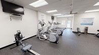 Located at 841 Gawain Cir in Dayton, Ohio, this fitness center is a well-equipped, single-floor facility designed to support a comprehensive range of workout routines. The space features durable rubber flooring throughout, providing a safe and functional surface for all types of exercise. Overhead, a suspended drop ceiling with recessed fluorescent lighting ensures the entire area is brightly and evenly illuminated.

The main workout area is organized to maximize usability. A row of Precor treadmills lines one wall, accompanied by Precor elliptical cross-trainers, offering users quality cardio options. A wall-mounted flat-screen television is positioned near the cardio equipment for entertainment during workouts. A dedicated strength training alcove houses a three-tier dumbbell rack stocked with a full range of hex dumbbells, alongside an adjustable weight bench — ideal for targeted resistance training. A multi-station cable machine adds further versatility for strength exercises.

For functional fitness, the space is stocked with a variety of exercise and medicine balls displayed on a tall storage rack, along with a rowing machine. Full-length mirrors along one wall expand the sense of space and allow users to monitor their form. Natural light filters in through double doors and windows, and the layout allows for comfortable circulation throughout. The facility is clearly marked with safety signage and emergency exits, reflecting a well-maintained and safety-conscious environment.