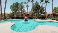 This outdoor amenity space showcases a generously sized freeform swimming pool as its centerpiece, highlighted by a distinctive rock-feature fountain that jets water from the middle of the pool. The pool's curvilinear design creates a resort-like aesthetic, with clear turquoise water and wide concrete decking surrounding the perimeter, offering ample space for sunbathing and leisure.

The deck is well-furnished with green lounge chairs and seating arrangements, complemented by large green patio umbrellas that provide shaded relief. A retractable striped awning extends over a dedicated seating area, adding an additional covered retreat for guests.

The space is framed by mature desert landscaping, including tall swaying palm trees and full-canopied pine trees, which lend a sense of privacy and natural enclosure. A secure perimeter fence defines the boundary of the pool area, while well-maintained shrubs and plantings add greenery throughout.

In the background, a Mediterranean-influenced clubhouse building with a red-tiled pitched roof and warm stucco exterior serves as an architectural focal point. The overall ambiance is that of a well-appointed, resort-caliber outdoor retreat designed for relaxation and recreation in a sunny, arid climate.
