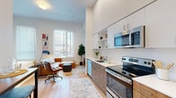 This efficiently designed single-floor unit offers a thoughtfully organized open-plan layout that maximizes every square foot. The main living area seamlessly integrates the kitchen, dining, and bedroom zones, creating a flexible and functional space. Soaring ceilings amplify the sense of openness, while large windows along the exterior wall flood the interior with generous natural light and provide outward views.

The kitchen is outfitted with stainless steel appliances, including an electric range, microwave, and dishwasher. Two-tone cabinetry pairs warm wood-finish lower cabinets with crisp white upper cabinets and open shelving, complemented by a clean white countertop and undermount sink. Light hardwood flooring runs throughout, enhancing the bright, airy atmosphere.

The primary living and sleeping area is generously proportioned relative to the unit's overall footprint, accommodating both a full bedroom setup and a comfortable lounge area. The full bathroom features a wood-tone vanity with a wide undermount sink, a large mirror, and a shower-tub combination. The overall design aesthetic is clean, contemporary, and efficiently planned.