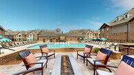 Located at 110 Singh Ln in Cary, North Carolina, this single-level outdoor amenity space offers a thoughtfully designed resort-style retreat. The centerpiece is a generously sized swimming pool framed by classic brick coping, surrounded by an expansive stone-tiled sundeck lined with lounge chairs and teal-accented umbrellas, providing ample space for relaxation under open skies.

Adjacent to the pool, a dedicated fire pit lounge area features a sleek linear gas fire pit set into a raised stone surround, flanked by cushioned wooden armchairs with warm accent pillows — an inviting gathering spot for cooler evenings. A poolside bar counter with high seating offers a casual social setting with direct views of the water.

The outdoor fireplace terrace is a standout architectural feature, showcasing a soaring natural stone chimney set against a rich brick exterior wall. Arched French doors with divided-light windows frame the fireplace on either side, while comfortable upholstered seating creates an intimate conversation area on the stone-paved patio.

The surrounding buildings display traditional brick and stone craftsmanship with pitched rooflines, dormer windows, and cohesive detailing throughout. The entire outdoor space benefits from abundant natural light and open sightlines, combining functional amenities with an elevated, community-oriented ambiance.