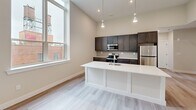 Located at 70 Church Street in Flemington, NJ, this thoughtfully designed 2-bedroom, 1-bathroom residence offers a clean, modern aesthetic across a well-appointed single floor. Light-toned luxury vinyl plank flooring flows seamlessly throughout, complementing the neutral palette of soft whites and warm greiges that define the interior.

The open-concept kitchen and living area serves as the heart of the home, anchored by a striking white kitchen island with an undermount sink. Dark espresso cabinetry lines the kitchen walls, paired with stainless steel appliances and sleek pendant lighting overhead. Tall windows flood the space with abundant natural light and provide views of the surrounding streetscape.

The generous primary bedroom features a spacious double-door closet with black hardware accents and soaring ceilings, creating an airy, comfortable retreat. The second bedroom also benefits from large windows with outdoor views and light-filled interiors. Three closets total provide ample storage throughout the unit.

The bathroom showcases a white shaker-style vanity with quartz countertop, a black-framed rectangular mirror, and modern matte black fixtures — a refined, contemporary touch. A dedicated laundry area and a well-lit hallway with a recessed access panel complete the functional layout. The dining room features additional cabinetry with gold-tone hardware and natural light from a nearby window.