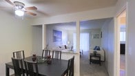 This single-floor space showcases a thoughtfully designed open-concept layout that seamlessly connects multiple living areas. The dining area serves as a welcoming entry point, featuring a dark-toned dining table and chairs set beneath a ceiling fan, with a textured ceiling that adds subtle character overhead. A low half-wall partition defines the boundary between the dining space and the main living room beyond, creating a sense of separation while preserving the open, airy feel throughout.

The central living room is bright and inviting, highlighted by a bold cityscape artwork that serves as a focal point on the accent wall. Vertical blinds along the far wall filter natural light generously into the space, contributing to the light and neutral ambiance. Plush carpeting in the living areas adds warmth underfoot.

A secondary sitting area is visible to the right, offering additional lounging space furnished with a white sofa and a sleek dark coffee table. The cohesive neutral color palette — soft whites, grays, and beige tones — flows consistently across all areas, creating a calm, unified atmosphere. Together, the connected spaces offer a functional and comfortable single-level layout well-suited for both everyday living and entertaining.