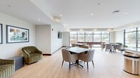 Located on the second floor at 1415 County Road B W in Roseville, Minnesota, this community room is a thoughtfully designed shared space that blends modern aesthetics with practical functionality. The open-concept layout seamlessly connects a fully equipped kitchen with a spacious lounge and dining area, creating an inviting environment ideal for gatherings and social interaction.

The kitchen is a standout feature, boasting sleek white cabinetry, a large center island with a quartz countertop, stainless steel appliances including a refrigerator and range, a stone slab backsplash, and a dedicated range hood. Floor-to-ceiling windows flood the kitchen with natural light while offering lush, treetop views of the surrounding landscape.

The open lounge and dining area is anchored by light hardwood flooring throughout and recessed ceiling lighting that creates a warm, welcoming ambiance. Multiple seating arrangements with contemporary upholstered chairs and pedestal tables provide flexible options for dining or conversation. A wall-mounted television and framed artwork add thoughtful finishing touches. Large windows along the perimeter frame scenic outdoor views and bathe the space in abundant natural light. A shuffleboard table adds a recreational element, rounding out this well-appointed community gathering space.