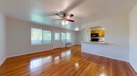 This well-maintained single-floor residence offers a clean, functional layout with warm hardwood flooring that runs consistently throughout the living spaces and bedrooms, lending the home a cohesive and inviting character. The expansive living area is bathed in natural light from a generous bank of windows fitted with blinds, offering views of lush greenery outside. A ceiling fan with an integrated light fixture provides both comfort and ambiance in the main living space. The kitchen is positioned adjacent to the living area and is partially open via a pass-through design, featuring warm wood cabinetry, granite-style countertops, a stainless steel sink, and black appliances including a microwave, range, and dishwasher. Tile flooring in the kitchen provides a practical contrast to the hardwood elsewhere. The bedrooms continue the hardwood flooring theme and are each equipped with ceiling fans and windows that allow natural light to filter in. One bedroom features a built-in closet with bifold doors. The bathroom is neatly appointed with a wood vanity, a wall mirror, tile-surround soaking tub, and light tile flooring. Overall, the space presents a tidy, functional, and comfortable living environment with consistent finishes throughout.