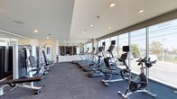 This fitness center offers a contemporary and well-appointed exercise environment designed to accommodate a wide range of workout needs. The space is characterized by expansive floor-to-ceiling windows along one wall, flooding the interior with natural light and providing open views of the surrounding exterior. Recessed lighting throughout the ceiling supplements natural light, ensuring consistent illumination across the entire facility.

The cardio zone is prominently featured, housing a row of treadmills equipped with individual digital display screens, alongside elliptical trainers and stationary bikes. The equipment is arranged to maximize the outdoor views, enhancing the exercise experience. A dedicated strength training area includes a well-organized dumbbell rack with a comprehensive range of weights, complemented by a large wall mirror that visually expands the space. Additional strength equipment includes a multi-functional cable machine stocked with various resistance accessories, medicine balls, and a stability ball.

The flooring consists of durable rubberized material suited for high-traffic fitness use. A separate mirrored section visible through glass partitions provides additional training space. The overall design is clean, modern, and functional, creating an efficient and motivating environment for users.