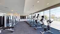 This fitness center offers a contemporary and well-appointed exercise environment designed to accommodate a wide range of workout needs. The space is characterized by expansive floor-to-ceiling windows along one wall, flooding the interior with natural light and providing open views of the surrounding exterior. Recessed lighting throughout the ceiling supplements natural light, ensuring consistent illumination across the entire facility.

The cardio zone is prominently featured, housing a row of treadmills equipped with individual digital display screens, alongside elliptical trainers and stationary bikes. The equipment is arranged to maximize the outdoor views, enhancing the exercise experience. A dedicated strength training area includes a well-organized dumbbell rack with a comprehensive range of weights, complemented by a large wall mirror that visually expands the space. Additional strength equipment includes a multi-functional cable machine stocked with various resistance accessories, medicine balls, and a stability ball.

The flooring consists of durable rubberized material suited for high-traffic fitness use. A separate mirrored section visible through glass partitions provides additional training space. The overall design is clean, modern, and functional, creating an efficient and motivating environment for users.