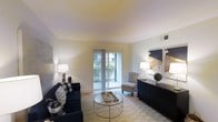This single-story space centers around a thoughtfully designed living room that balances comfort and style. The room is finished with smooth, light-colored walls and a clean white ceiling that helps amplify the sense of openness throughout. A large sliding glass door serves as the focal point of the space, flooding the interior with natural light and providing direct access to a private balcony that overlooks surrounding greenery and neighboring architecture.

The living area is furnished with a deep navy blue sofa accented by black-and-white plaid throw pillows, paired with a coordinating houndstooth accent chair. A circular glass-and-gold coffee table anchors the seating arrangement atop a textured area rug, adding a touch of contemporary elegance. To one side, a dark wood dresser is styled with decorative accents including a small topiary and table lamp, while a framed abstract artwork adds visual interest to the wall above.

Warm ambient lighting from table lamps on both sides of the room complements the natural light, creating a balanced and inviting atmosphere. The overall aesthetic is modern yet approachable, with a neutral palette punctuated by bold accent pieces.
