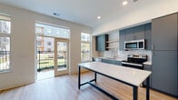 This single-floor unit showcases a clean, contemporary design with an open-concept layout that seamlessly connects the kitchen and living area. Light wood-tone flooring runs throughout, complementing the crisp white walls and recessed lighting that keep the space bright and welcoming.

The kitchen is a standout feature, outfitted with slate-blue flat-front cabinetry extending to the ceiling, a white subway tile backsplash, and a full suite of stainless steel appliances including a refrigerator, range, microwave, and dishwasher. Open wood shelving adds a modern touch, while a large island with a white countertop and bold black metal frame provides additional prep space and casual seating.

The living area benefits from abundant natural light streaming through large windows and a glass door that opens onto a private balcony, offering outdoor views of the surrounding community. A hallway leads to the additional rooms, including a bathroom finished with large-format tile flooring, a grey vanity with a white countertop, a full-width mirror, and a walk-in shower. A ceiling fan is also present in one section of the unit, adding to overall comfort. The thoughtful layout and modern finishes make this a well-appointed and functional living space.
