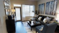 This single-floor space presents a thoughtfully designed living area that balances comfort and contemporary style. The living room serves as the centerpiece of the space, featuring rich dark hardwood flooring that grounds the room with warmth and elegance. Large windows dressed with white sheer curtains and horizontal blinds flood the space with natural light, creating a bright and welcoming atmosphere. A glass-paneled door provides direct access to the outdoors, further enhancing the sense of openness. The seating area is anchored by a plush dark charcoal sectional sofa accented with an array of decorative pillows in navy, white, and silver tones. A sleek dark wood coffee table sits atop a contemporary area rug, tying the seating arrangement together. Tasteful décor elements, including candlestick holders, a ceramic vase, and lush indoor greenery, add layers of visual interest throughout the room. Bold wall art featuring a nature-inspired print lends a dynamic focal point to the space. Ambient lighting from a classic table lamp complements the natural light, ensuring the room remains inviting at any time of day. The overall ambiance is modern, polished, and meticulously curated.