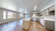 This single-story, 3-bedroom, 2-bathroom home features a thoughtfully designed open floor plan with consistent wood-style flooring flowing throughout every room. The combined kitchen and living area serves as the heart of the home, offering generous open space ideal for both everyday living and entertaining. The kitchen is well-appointed with gray shaker-style cabinetry, white appliances including a range and dishwasher, a ceiling-mounted range hood, a undermount sink, and a central island with a light countertop that provides additional prep and seating space. Multiple windows in the adjacent living area allow natural light to fill the space, and a ceiling fan with light kit promotes comfortable airflow. Each of the three bedrooms features wood-style flooring, a ceiling fan, and windows with outdoor views. The primary bedroom is a comfortable size and connects to a walk-in closet equipped with wire shelving systems for organized storage. A second closet provides additional storage elsewhere in the home. Both bathrooms are finished with gray vanity cabinetry, light stone-style countertops, large mirrors, and wood-style flooring. A hallway connects the bedroom wing to the main living areas, completing the home's efficient and functional layout.